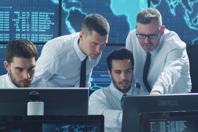 Pantalla digital, hombres vestidos de blanco y corbata negra observando monitores de computadoras