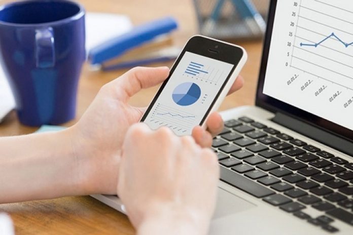 Lapto mostrando gráfico, manos sosteniendo celular, taza azul