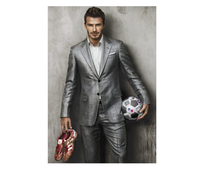 Hombre posando con traje gris, sosteniendo zapatos deportivos rojos y balón de fútbol de colores