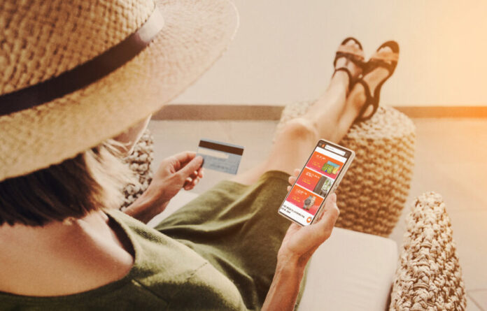 Mujer de piernas cruzadas, sombrero, en las manos celular mostrando aplicación y tarjeta de banco