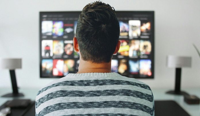 Hombre con franela de raya, parado al frente de un televisor mostrando aplicaciones