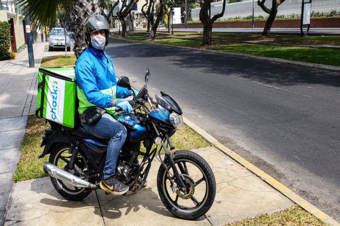 Calle, plaza, auto, hombre con casco, tapaboca, sentado en una moto azul y cargando un morral de envío con publicidad chazki