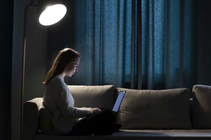 Mujer sentada sobre sofá observando laptop en habitación con cortinas y lámpara