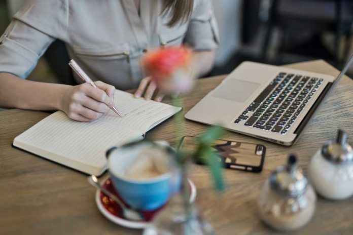 Mesa de madera, laptop, celular, taza de café, persona escribiendo en agenda