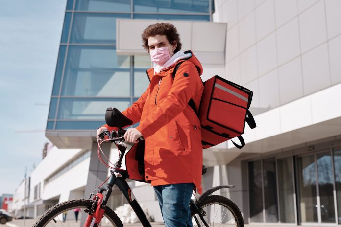 Hombre con tapaboca, chaqueta anaranjada en bicicleta con caja para delivery