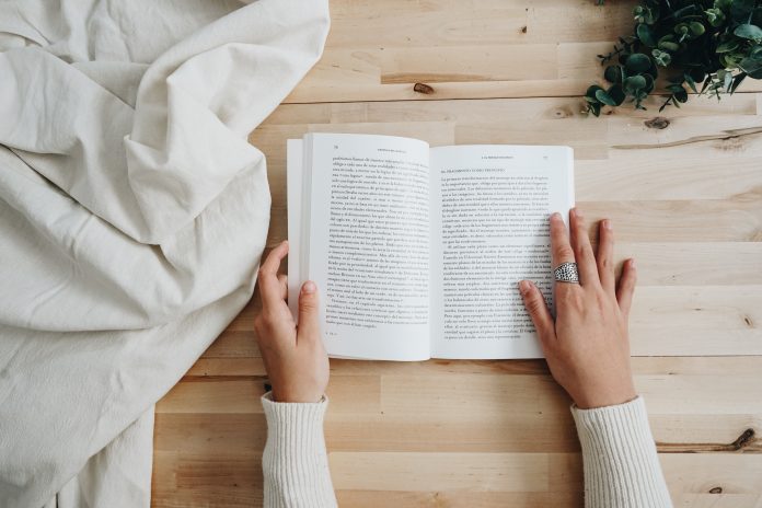 Manos de una persona con un suéter blanco leyendo un libro puesto sobre una mesa de madera