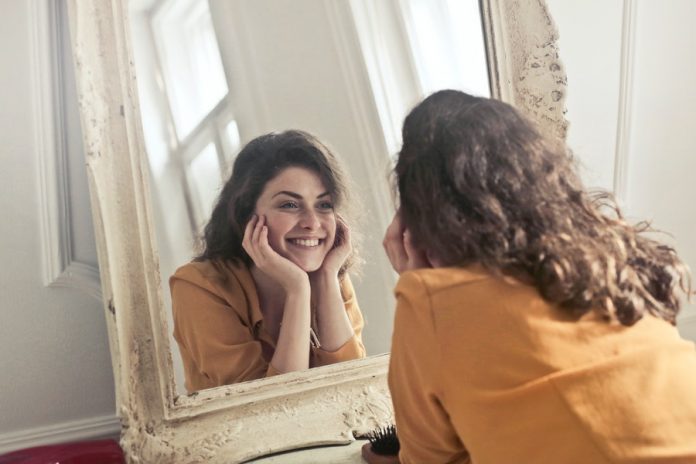 Mujer sonriendo, con las manos en la cara, vestida de amarillo, viéndose en un espejo