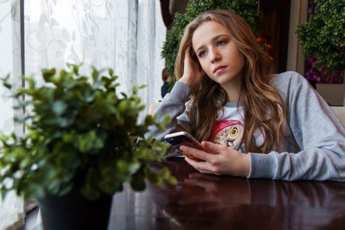 Adolescente con celular en la mano mirando por la ventana en una habitación con matas