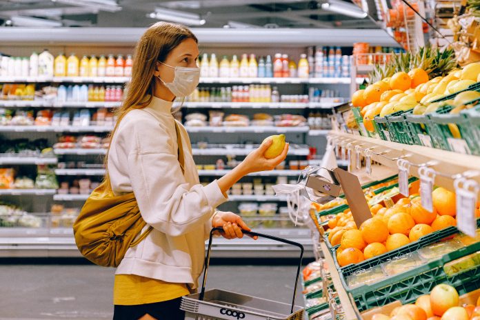 Mujer con tapaboca, bolso amarillo y cesta en las manos, en interior de supermercado con anaqueles y estantes llenos de productos
