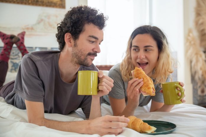 Personas acostadas en una cama, comiendo pan y tomando café