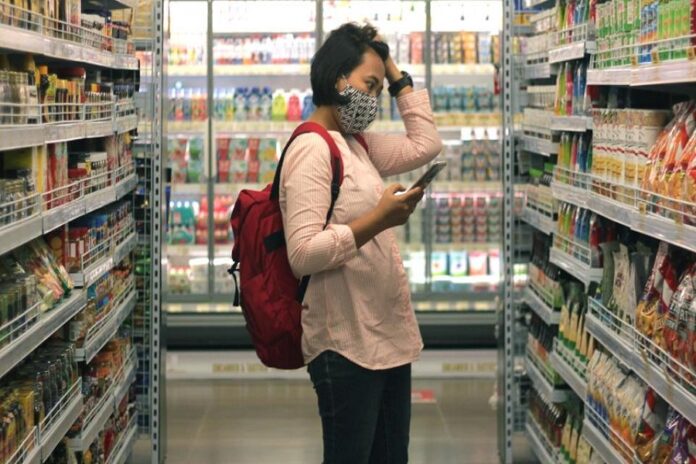 Interior de supermercado, anaquel y estantería con productos, mujer con celular y mano en la cabeza