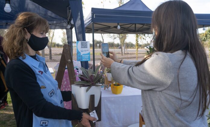 Mujer con celular en las manos capturando código Qr para una compra, vendedora con planta en las manos