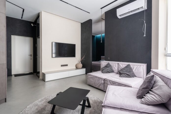 Interior de habitación, pared con estante y televisor, sofás con cojines, mesa negra y aire acondicionado en la pared